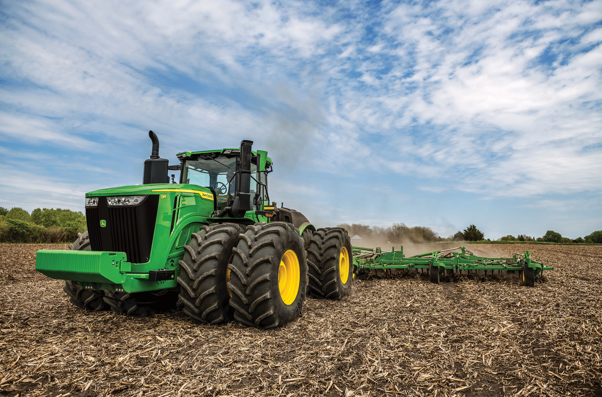 Green tractor with oversized dual tires pulling a cultivator across a harvested field with dust trailing behind the equipment under a cloudy blue sky