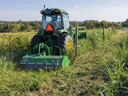 Rear view of tractor mowing tall grass along a fence line with a rear‑mounted mower attachment.