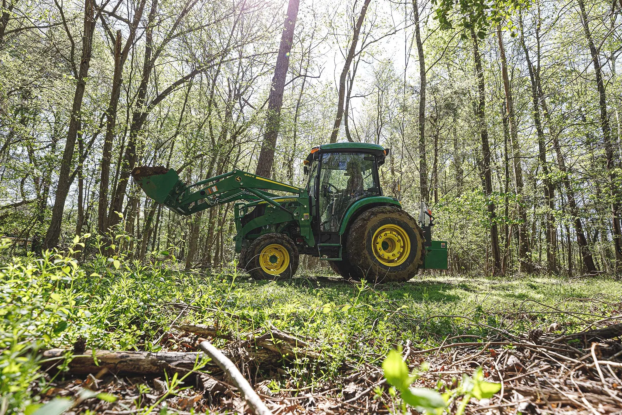 Side left view of a 4044R Compact Utility Tractor driving through a forest with loader extended, carrying a full load of dirt.