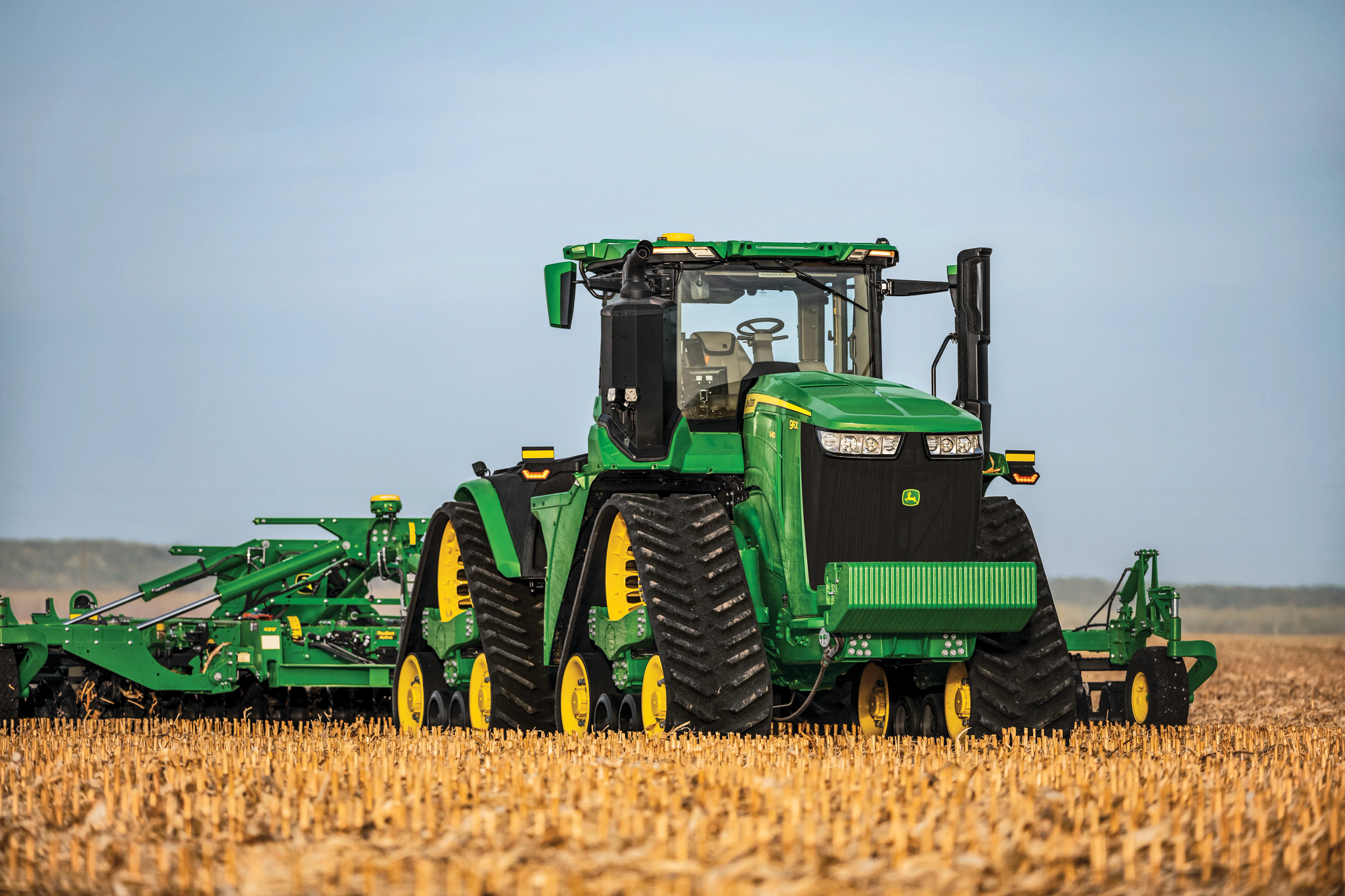 Front view of a large green tracked tractor with a tillage implement hitched to the rear sitting in a harvested field covered with corn stubble under a clear blue sky