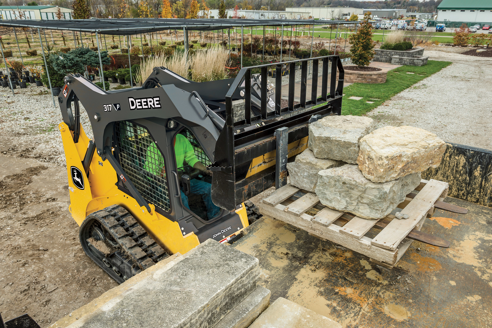 A compact track loader using a pallet fork to lift a wooden pallet stacked with large stone blocks in a landscaping yard