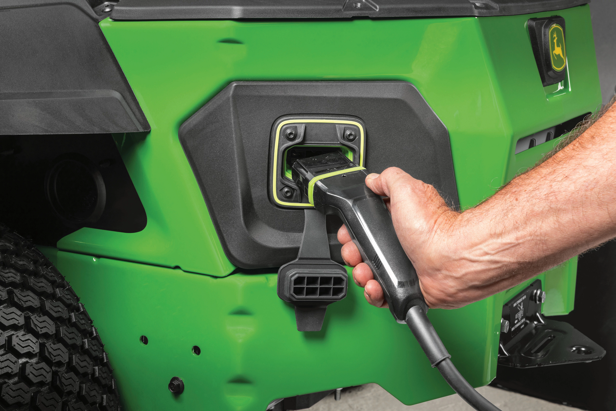 Close-up of John Deere equipment with a charging cable plugged into the side charging port; green body panel, Deere logo nearby, and a hand holding the connector.