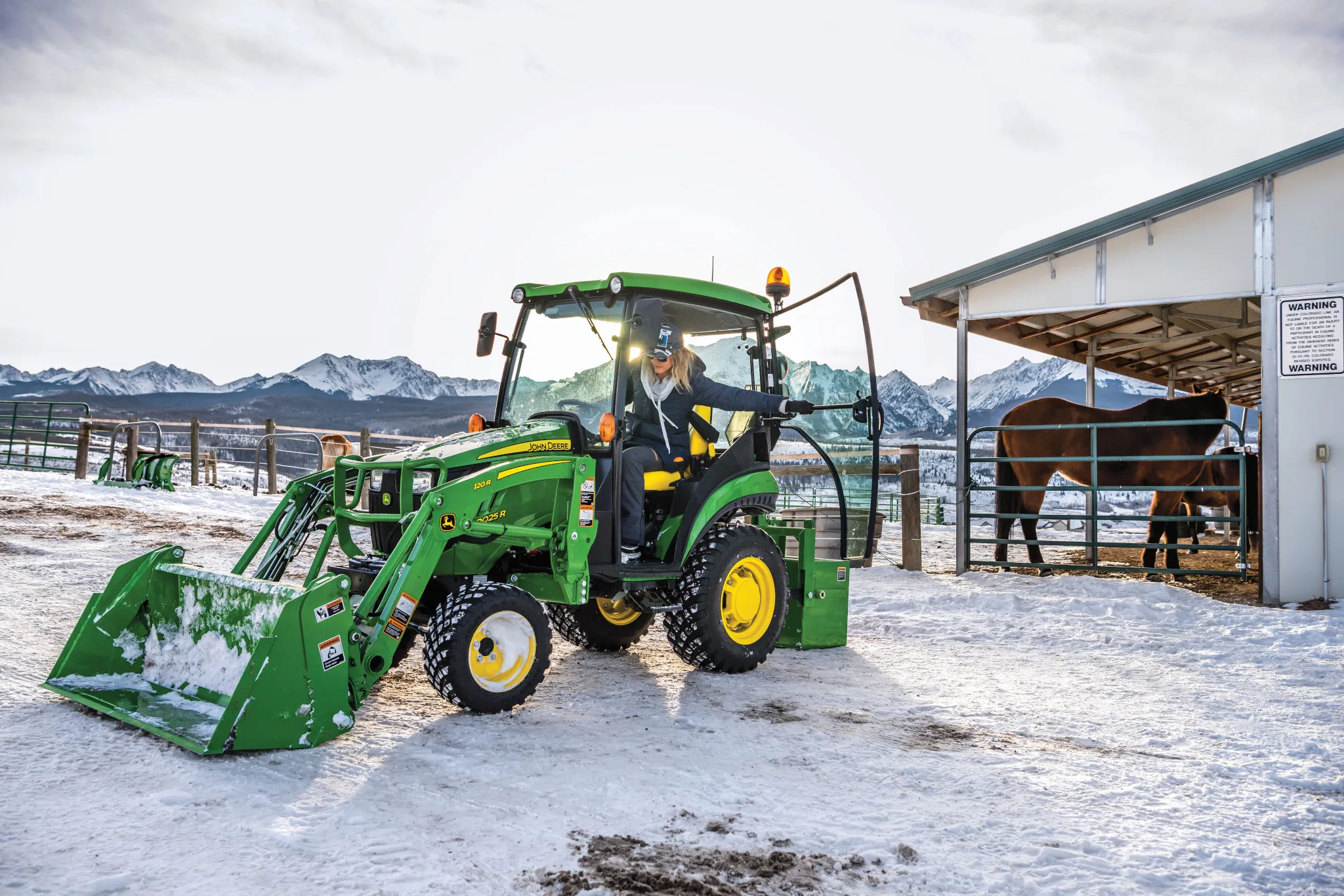 Green tractor with a front loader sitting on a snowy farmyard, as an operator steps out near a barn and fenced livestock area under a cloudy winter sky