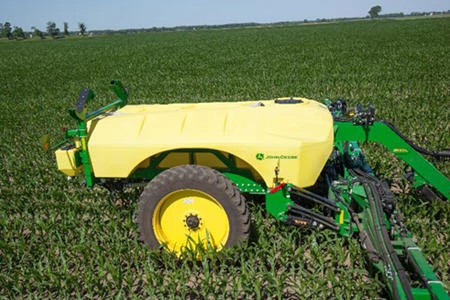 A liquid applicator with a large yellow tank and single rear wheel positioned in a green row-crop field with the green frame extended outward under a clear sky