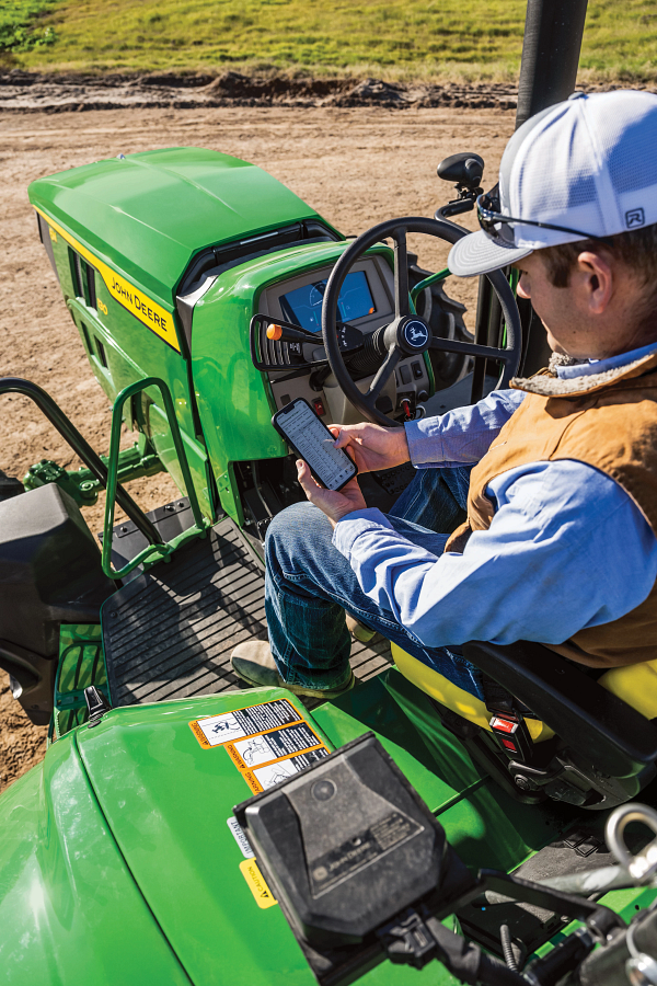 Operator seated on an open‑station John Deere tractor, viewed from above, holding a smartphone near the steering wheel; green hood, control levers, warning decals, and dirt roadway visible in an outdoor work setting.