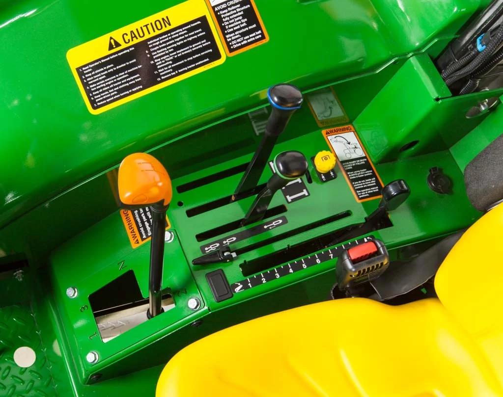 Close-up of John Deere tractor operator controls showing green console with labeled control levers, throttle and hydraulic controls, caution decals, and yellow seat edge, highlighting the machine’s operator station.