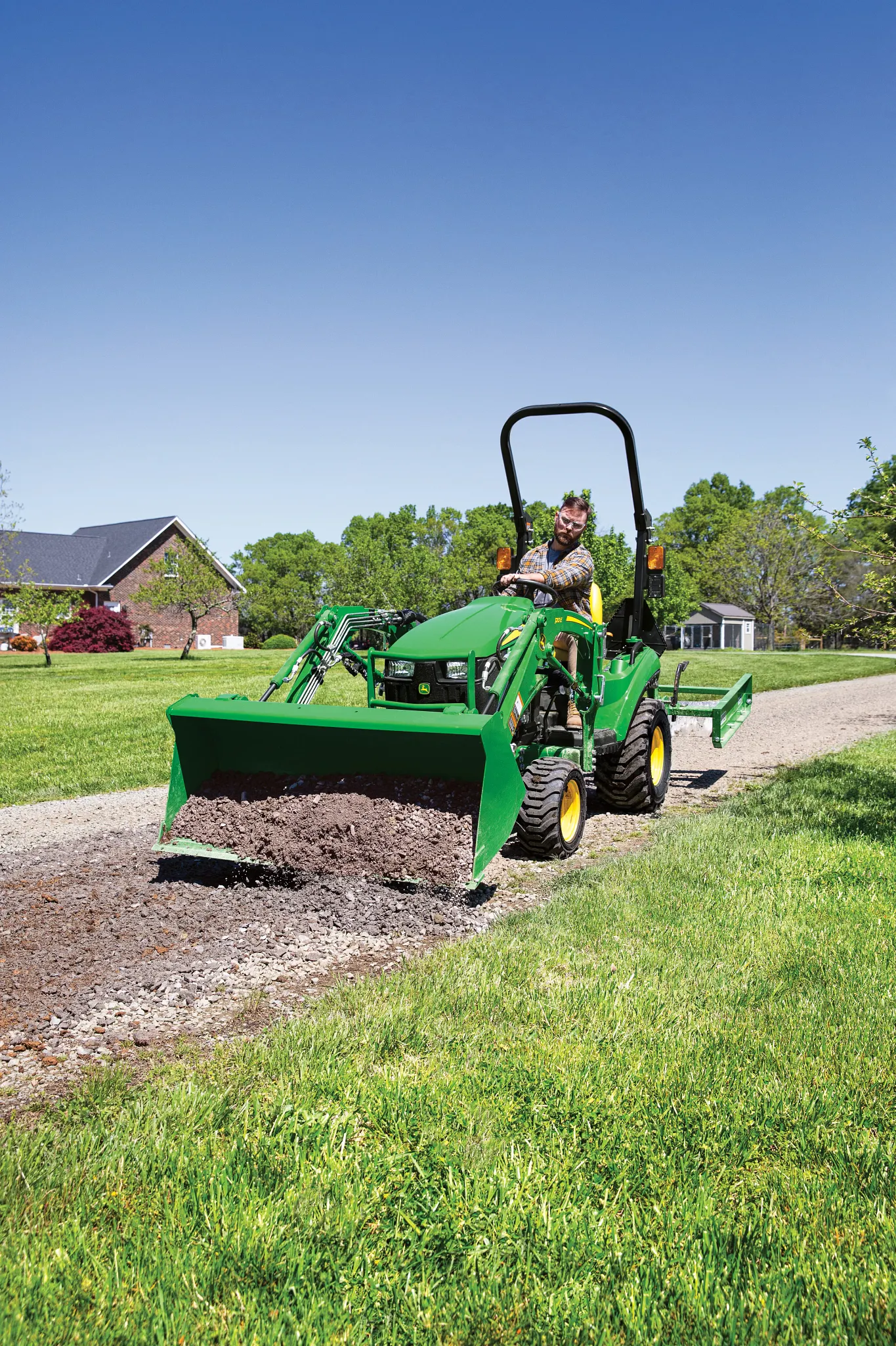 A person operates a small green tractor with a front loader attachment, leveling and spreading gravel along a driveway beside a grassy lawn, with houses and trees visible under a clear blue sky