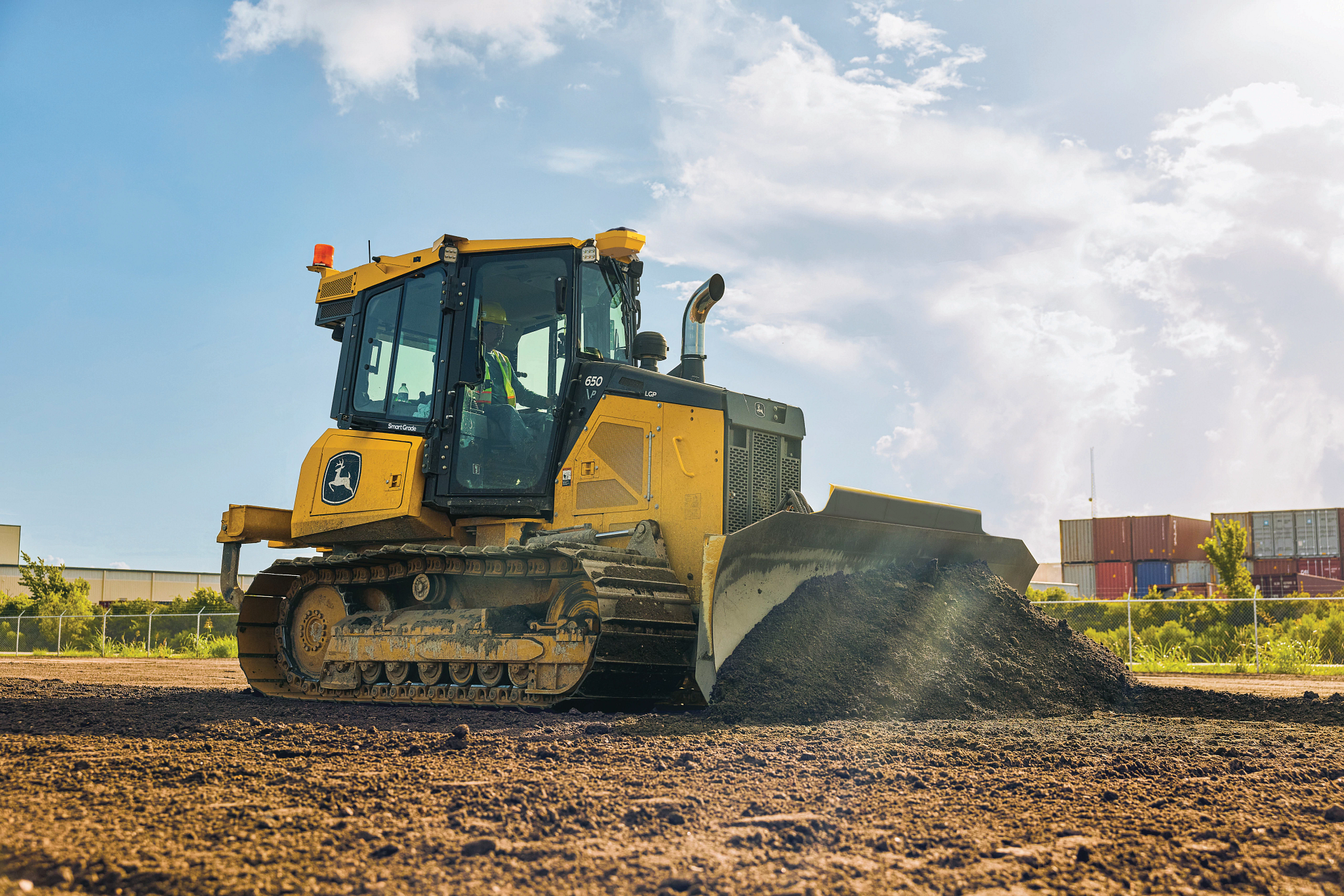 A yellow dozer with large blade, enclosed cab, and wide tracks pushing a pile of soil across an open construction site under a partly cloudy sky