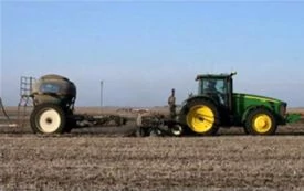 Wide shot of a green tractor pulling farm equipment and a large wheeled tank across a tilled field under a clear blue sky