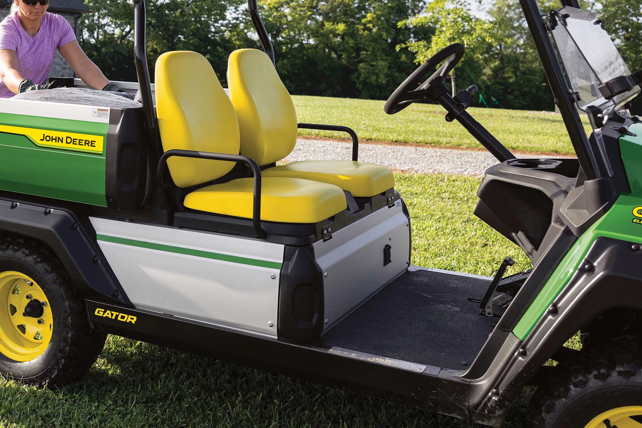 Close-up, side right view of a GX Electric Gator's operator platform and yellow seats. It is parked in the grass, and a landscaper is reaching for material in the cargo box.