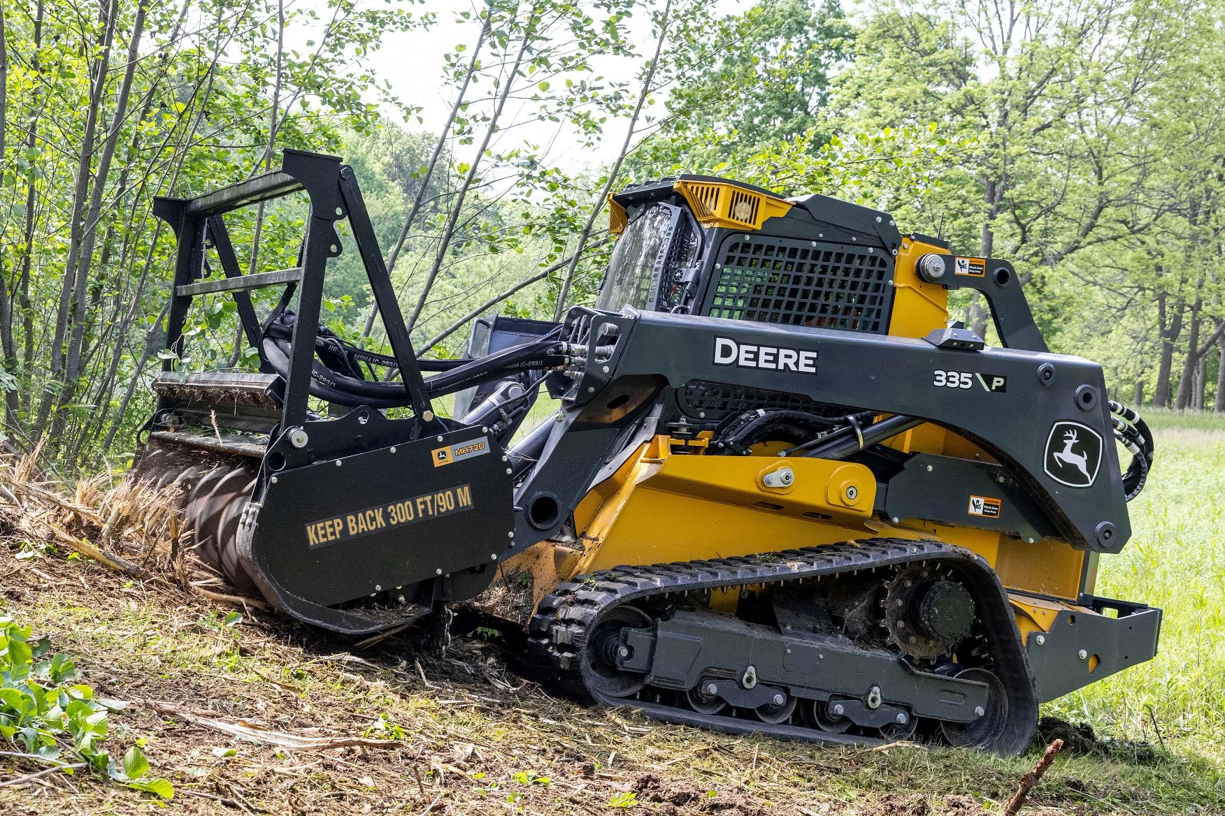 A yellow John Deere skid steer uses a front-mounted forestry mulcher attachment to grind brush and small trees in a wooded clearing.