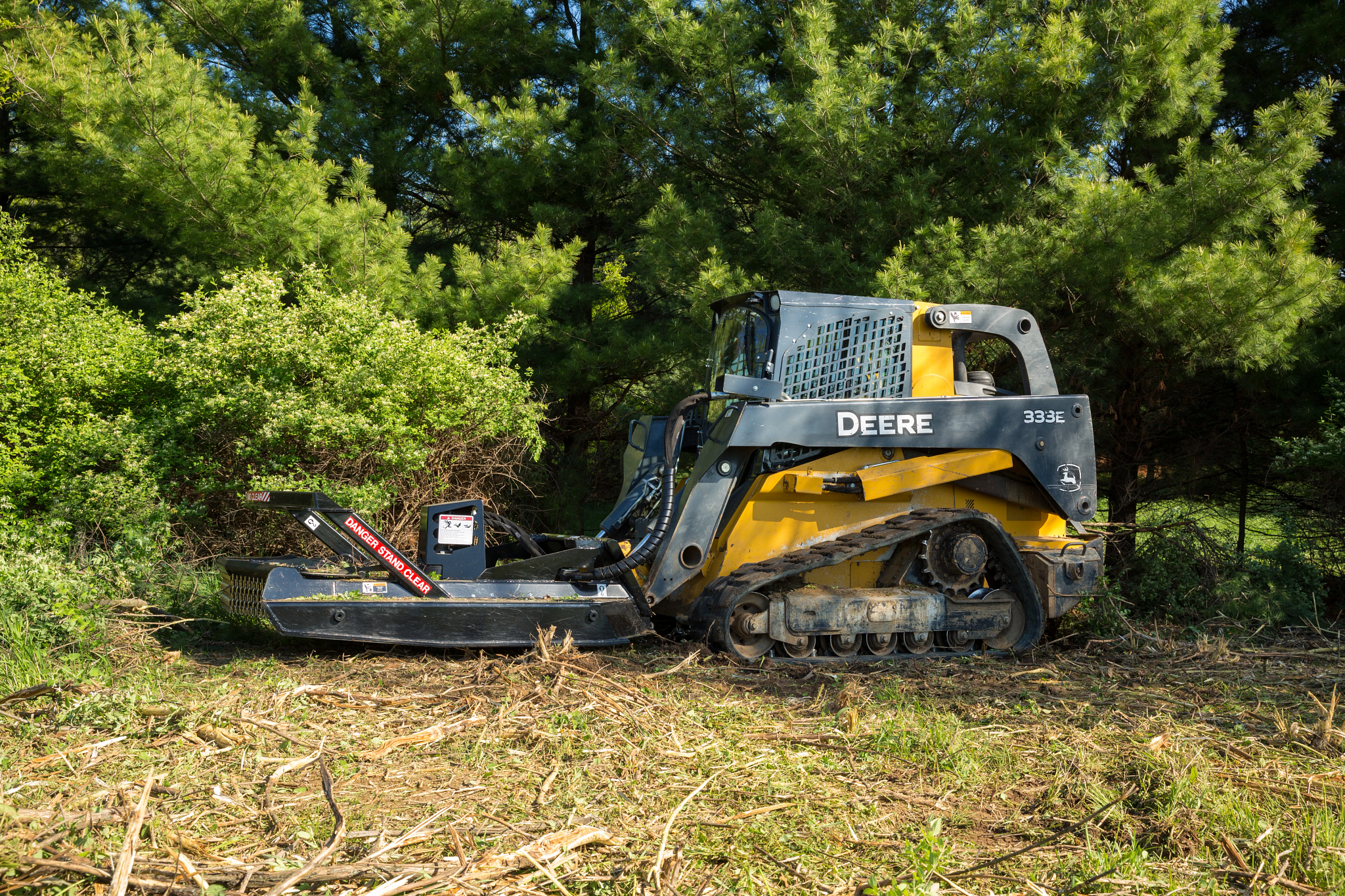333E Compact Track Loader with an RS72 Rotary Cutter clearing brush along a tree line