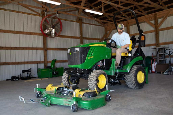John Deere 1025R sub‑compact tractor with mid‑mount mower deck parked inside a barn, shown with yellow wheels, ROPS, and operator seat, mower deck lowered on concrete floor.