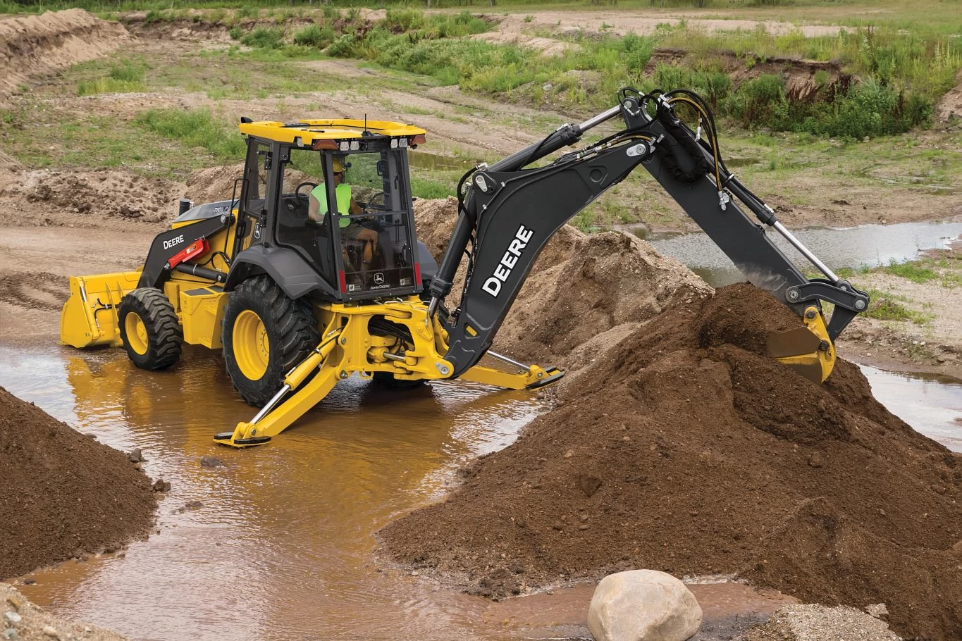 A yellow John Deere backhoe loader scoops a pile of brown soil beside shallow muddy water at a construction site.