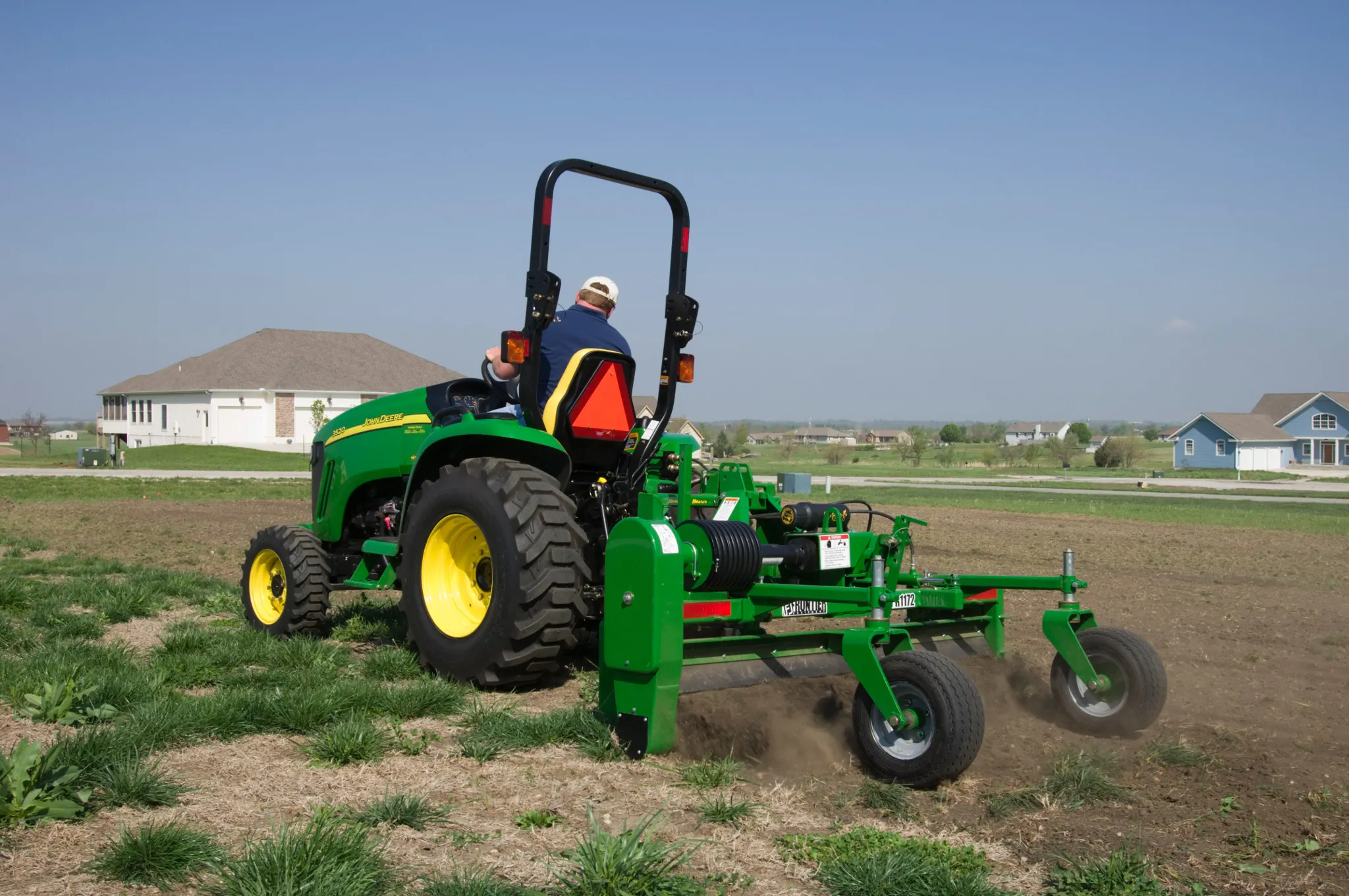 A person operating a green tractor with a rear-mounted soil cultivation attachment, moving across a dry, grassy field. Dirt is being turned behind the implement, with houses and open land visible in the background under a clear sky.