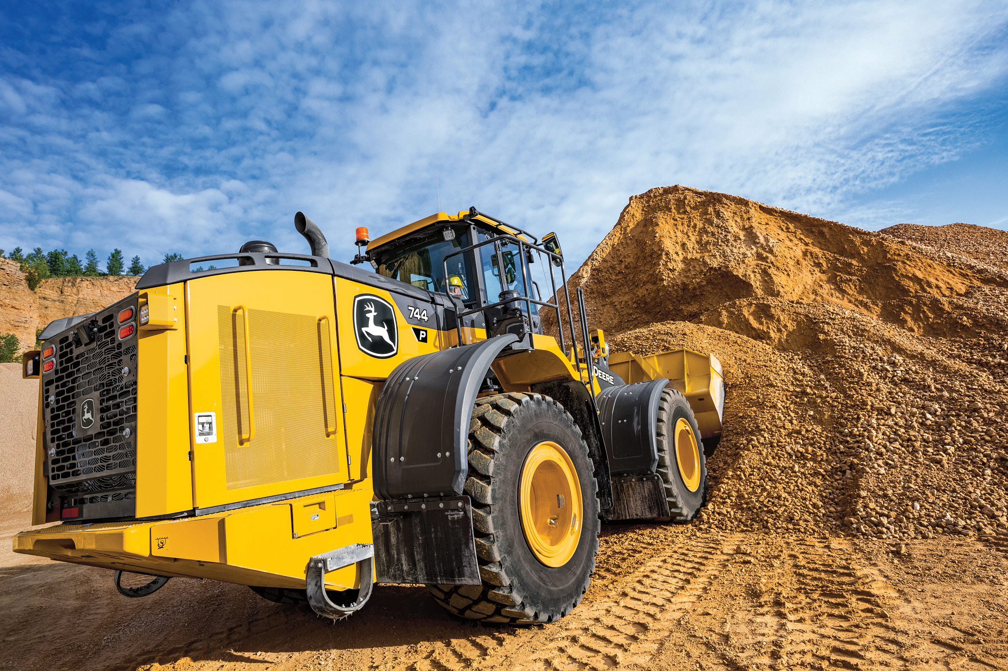 A large yellow front-end loader is positioned at the base of a tall gravel pile in a quarry, with its bucket partially buried in the mound