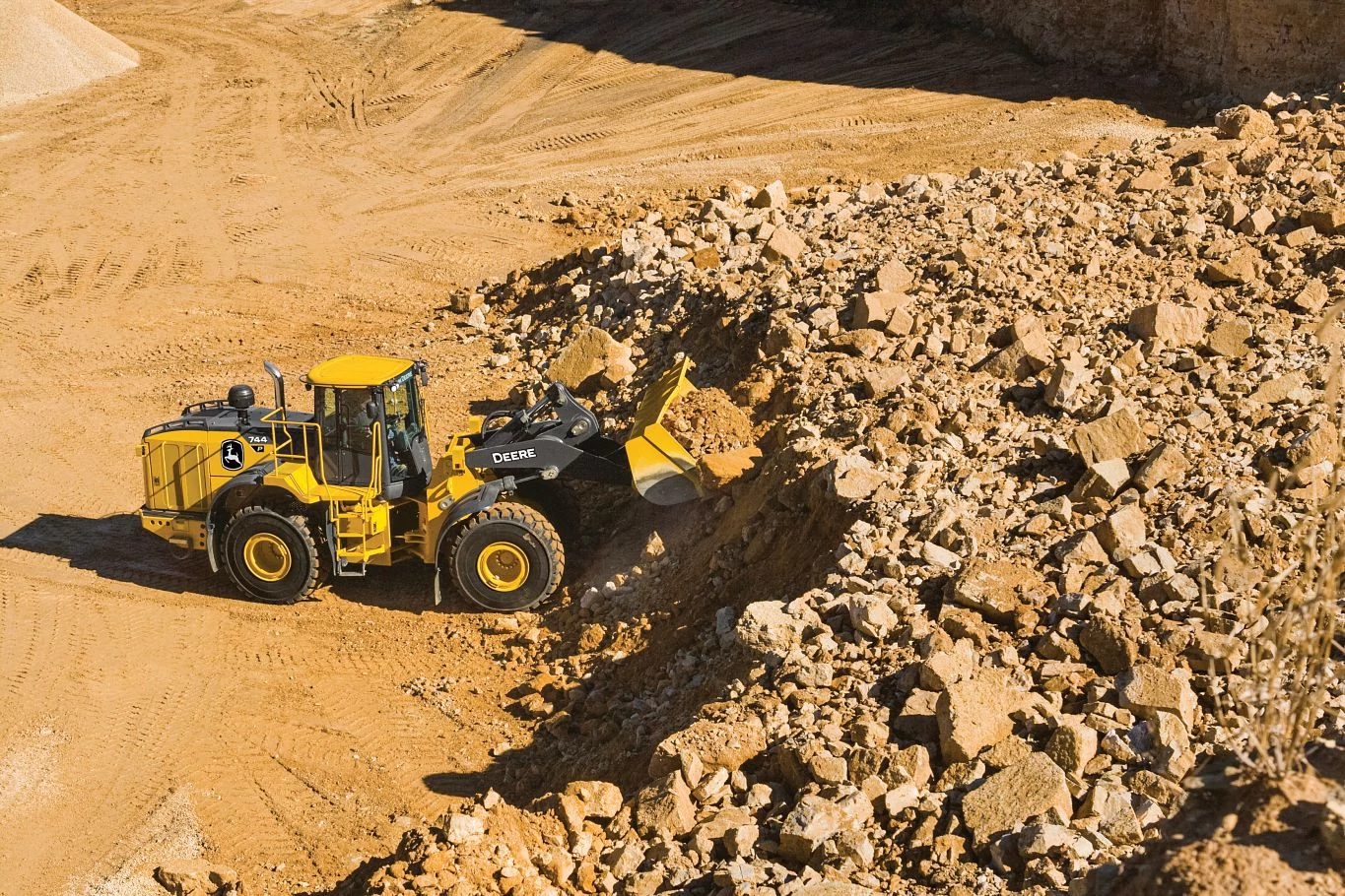 A yellow front-end loader operates in a quarry, using its bucket to scoop into a large pile of broken rocks and gravel