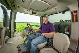 Interior cab view of a machine in a green field from the passenger's side door. A farmer with purple shirt and ballcap is steering.
