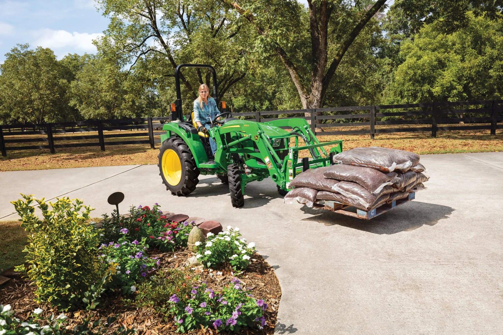 A green tractor with a front loader carries a pallet of bagged mulch across a paved driveway, with an operator seated at the controls, landscaped flowers in the foreground, and trees and fencing in the background