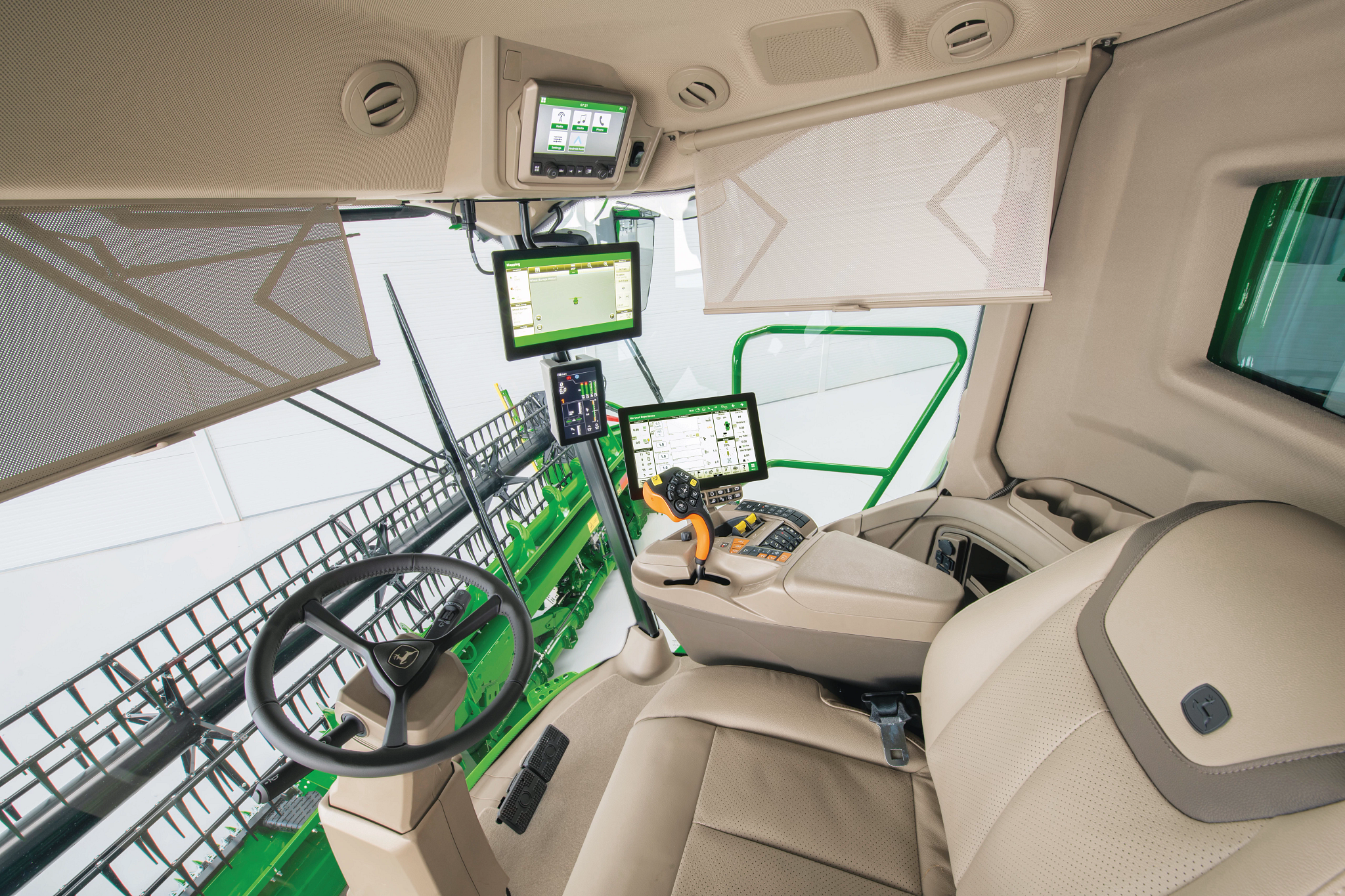 Interior of a combine cab showing a padded seat, steering wheel, multiple digital display screens, control consoles, and large windows with sunshades