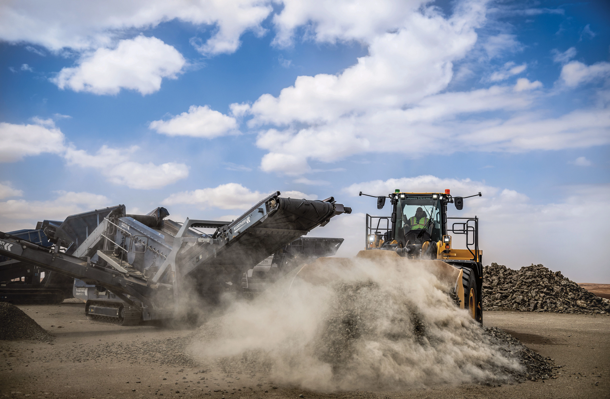 Front-facing wheel loader pushing a pile of gravel across a dusty work site, kicking up a cloud of dust, with a large rock-processing conveyor machine operating in the background under a partly cloudy sky