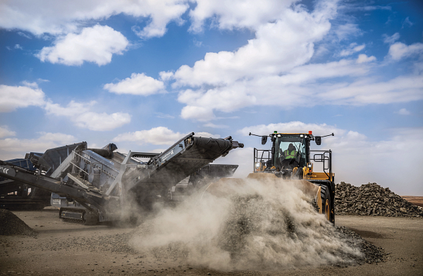 Front-facing wheel loader pushing a pile of gravel across a dusty work site, kicking up a cloud of dust, with a large rock-processing conveyor machine operating in the background under a partly cloudy sky