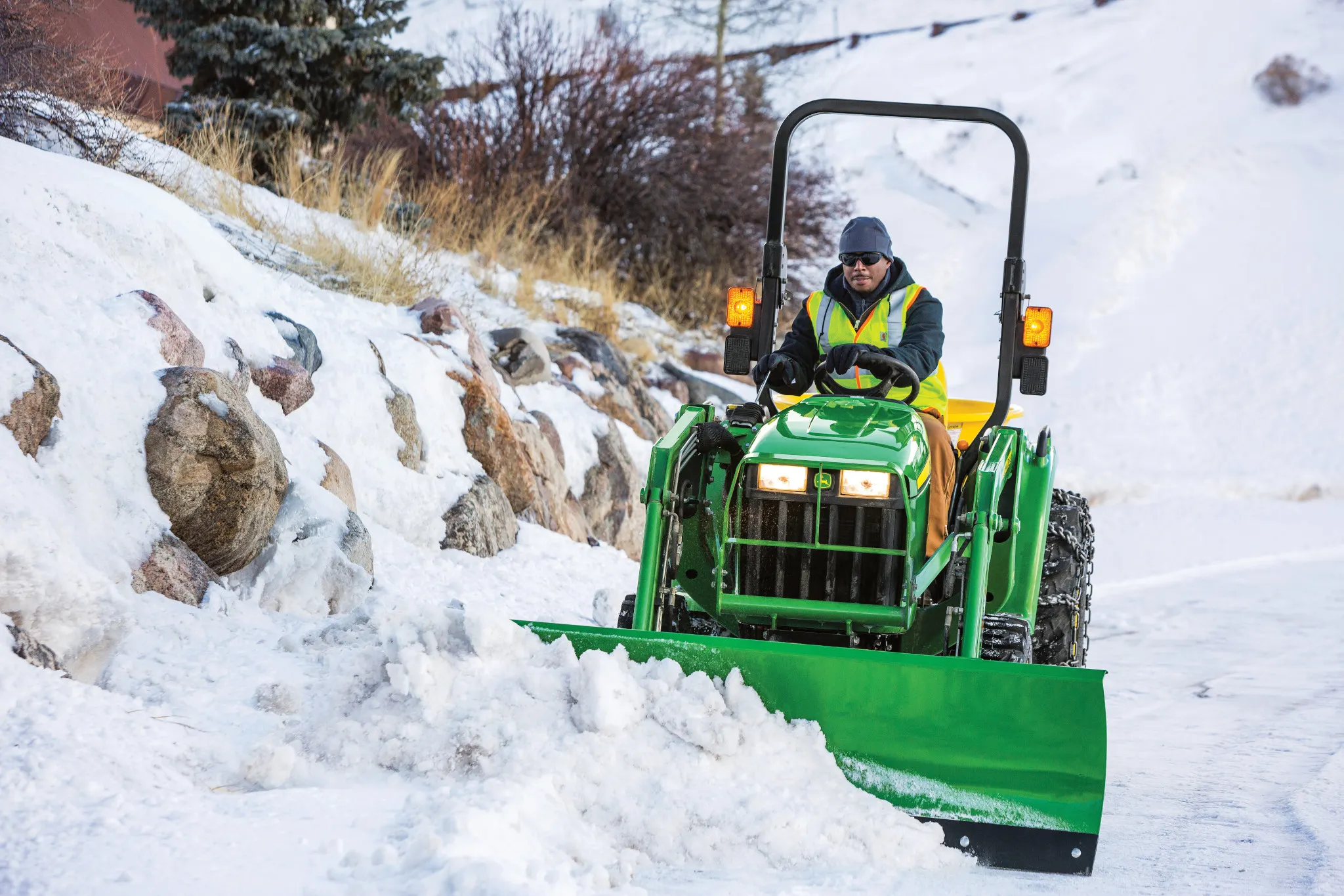 A small green tractor with a front-mounted plow clears a snow-covered road, driven by an operator wearing a high-visibility jacket, with rocks and grass lining the snowy roadside