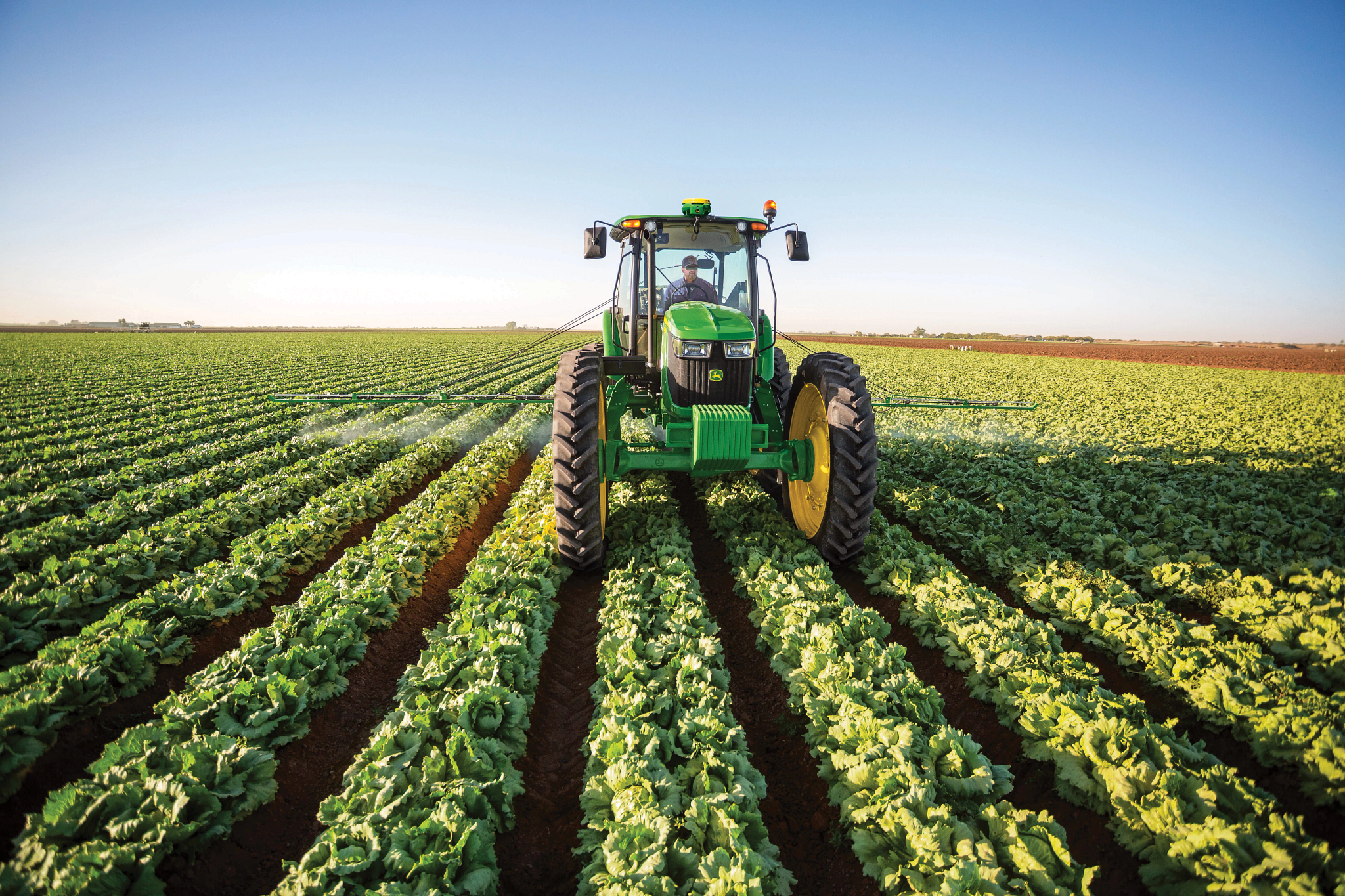 Front view of John Deere row‑crop tractor driving between rows of leafy green crops, with large front tires, enclosed cab, and clear blue sky over a cultivated field.