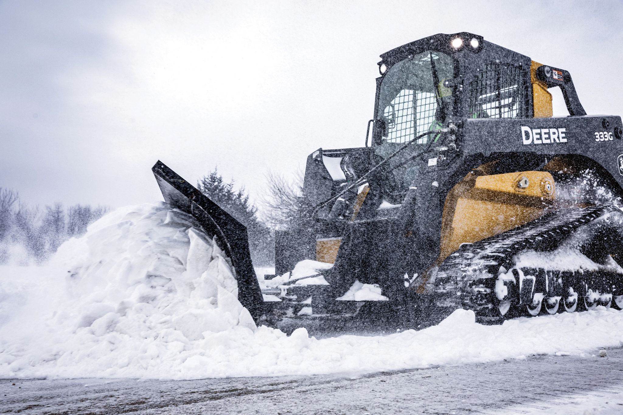 Side view of a tracked John Deere skid steer pushing a large pile of snow with a front-mounted blade on a snowy landscape