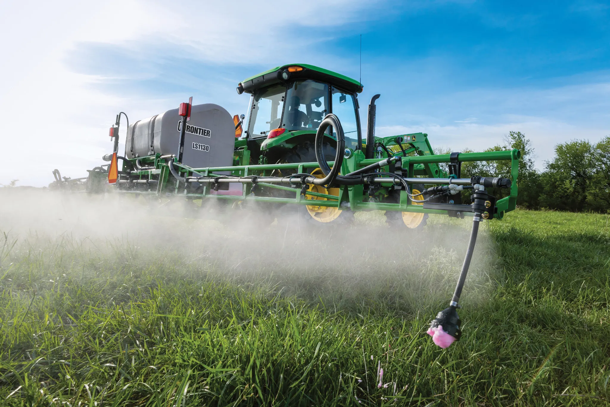 A agricultural sprayer tractor applies a fine mist over a grassy field using extended spray booms, with nozzles releasing chemicals close to the ground under a blue sky.