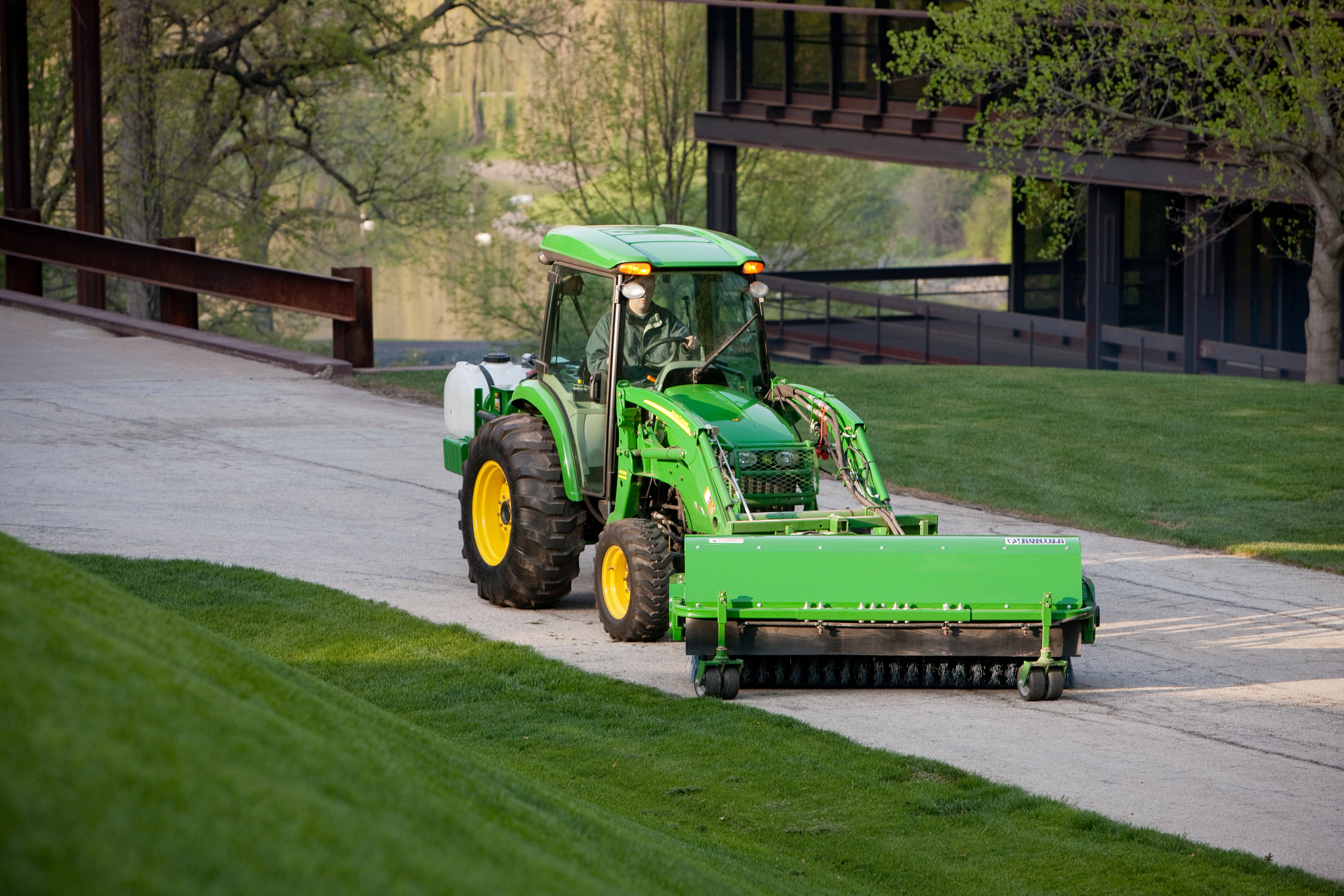 Front right view of a compact utility tractor angling a SW21 Series front mount rotary broom along a road between lawns