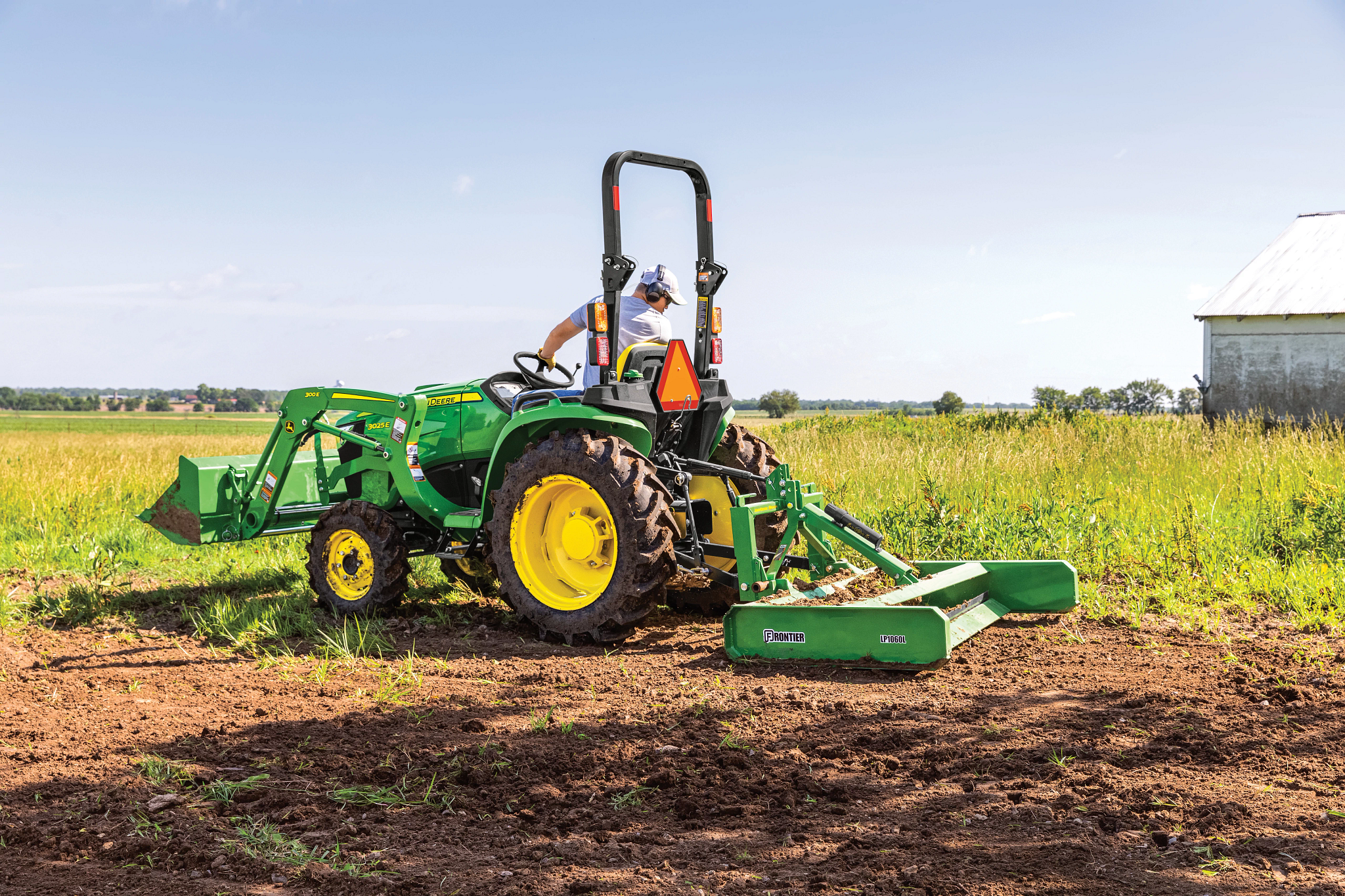 John Deere compact tractor with front loader and rear rotary cutter mowing a grassy field, operator seated under ROPS, yellow wheels and green body visible, with a barn and open farmland in the background.