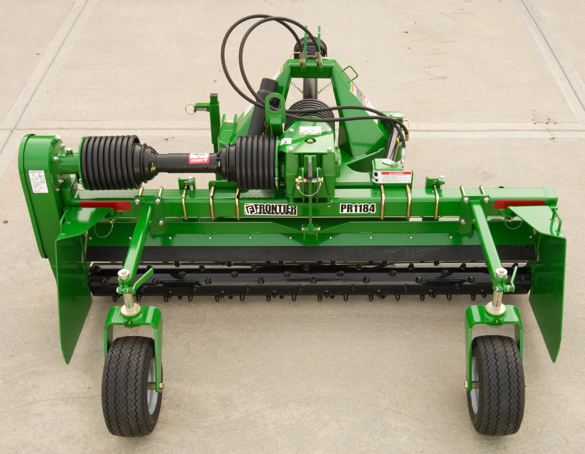 Frontier PR1184 rotary tiller attachment on concrete, featuring a wide cutting deck, central gearbox with PTO shaft, rows of blades underneath, and two support wheels.