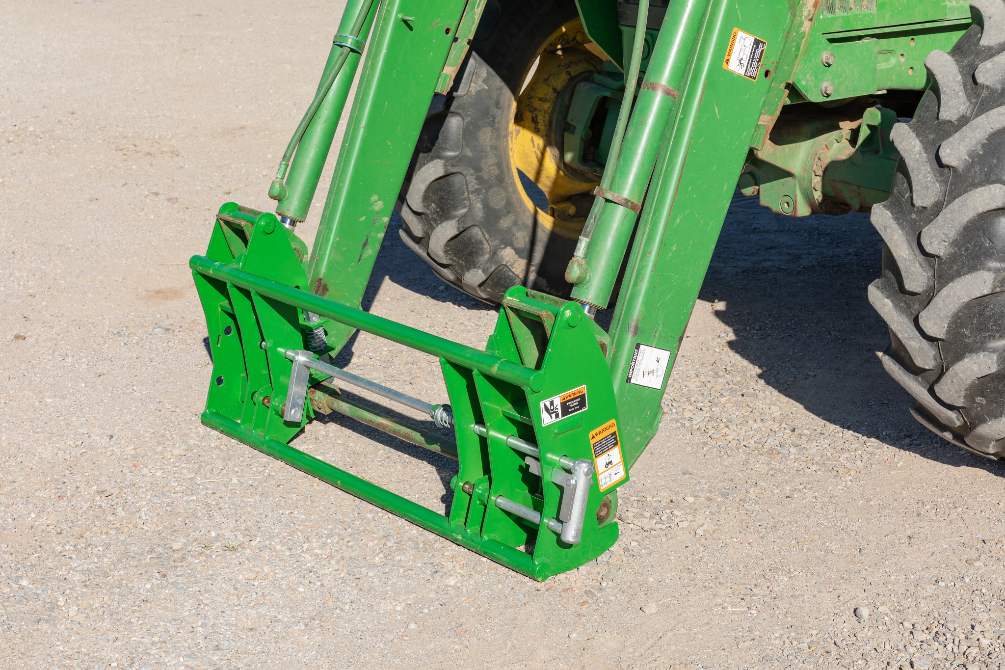 Close-up of a tractor front-loader mounting frame with metal linkage bars and hydraulic arms resting on a gravel surface, positioned between the tractor’s front wheels.