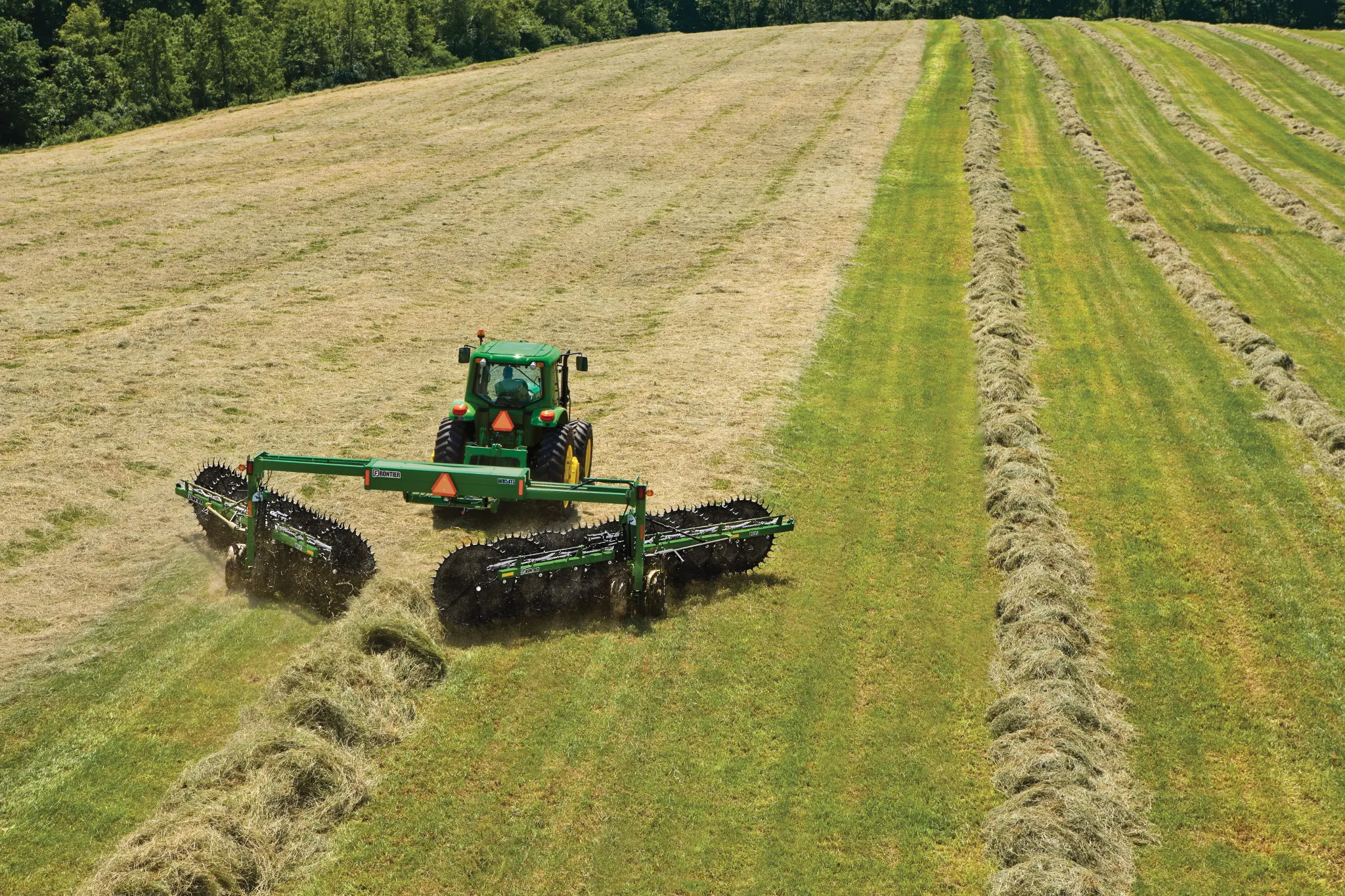 Rear aerial view of a tractor using a WR5417 wheel rake to form windrows of hay in a green field