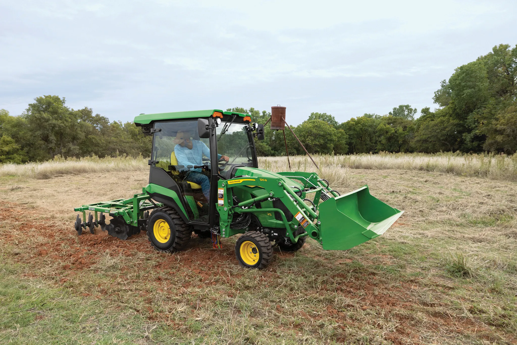 A green tractor with an enclosed cab and front loader operates in a field, pulling a disk harrow implement through dry grass and soil with trees and open land in the background