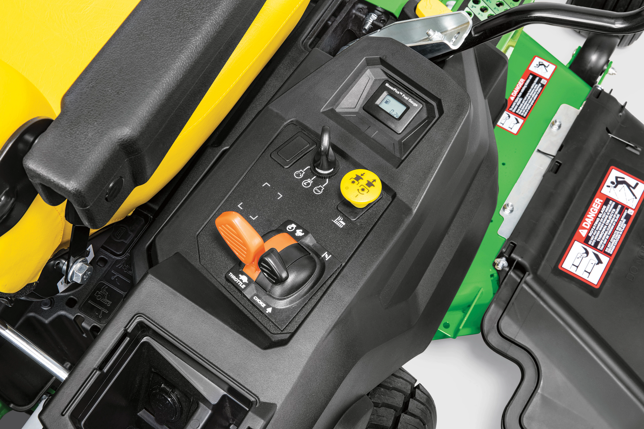 Close-up of a John Deere zero‑turn mower control console showing throttle and choke controls, PTO engagement knob, ignition switch, and warning labels mounted beside the yellow seat and green frame.