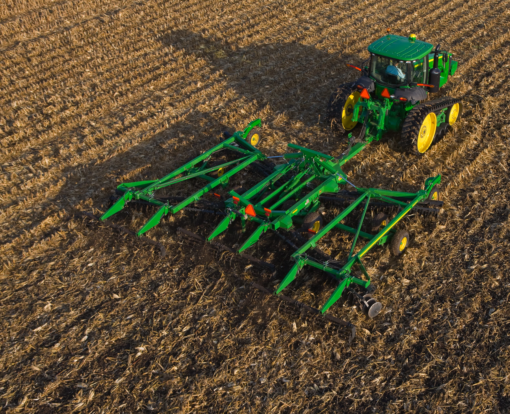 Aerial view of tractor pulling a wide green tillage implement across a harvested field, with soil residue visible and the equipment actively working the ground in parallel rows.