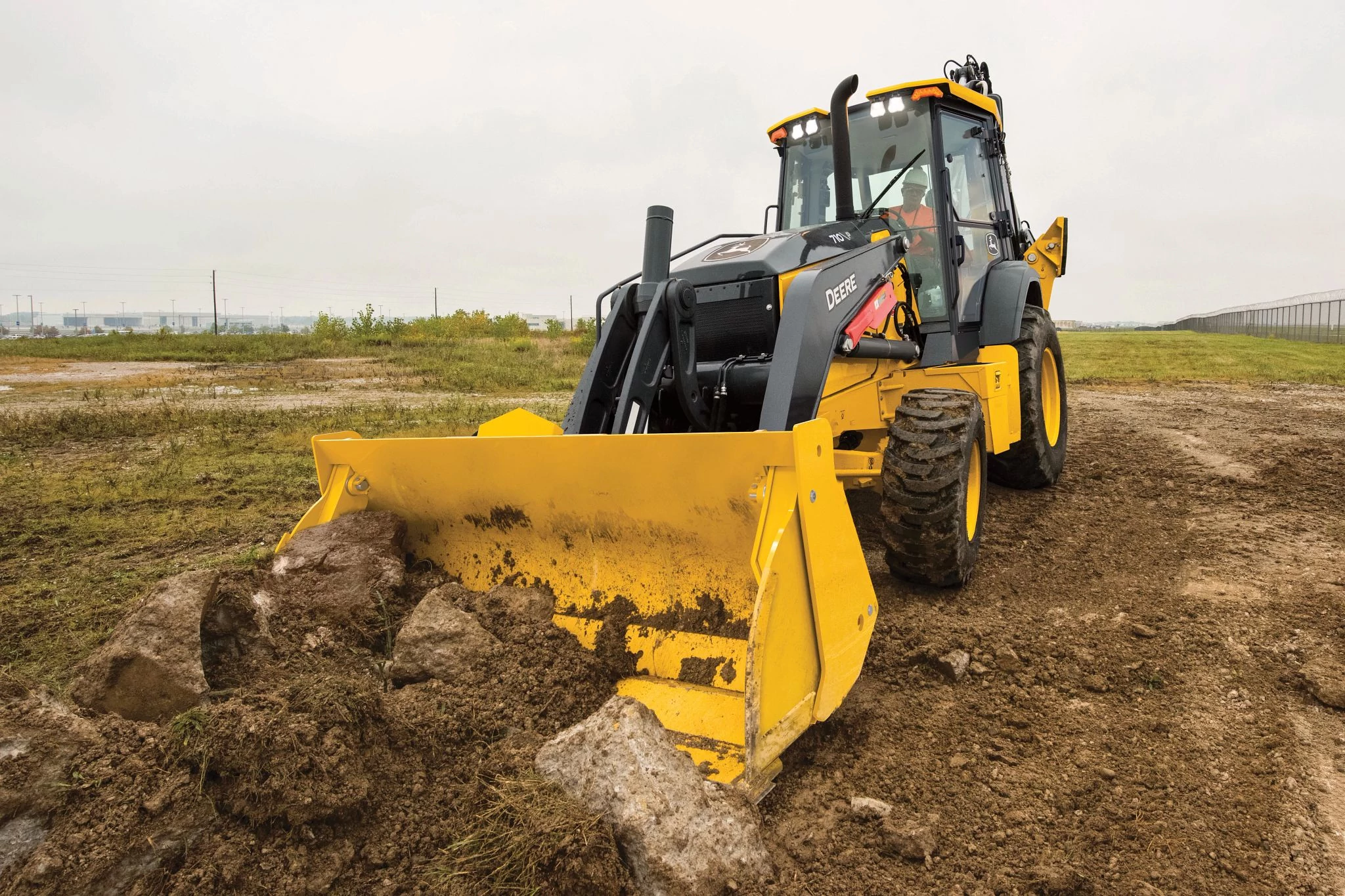 A yellow backhoe loader moves across a rough, muddy construction site, using its wide front bucket to push rocks and clumps of soil