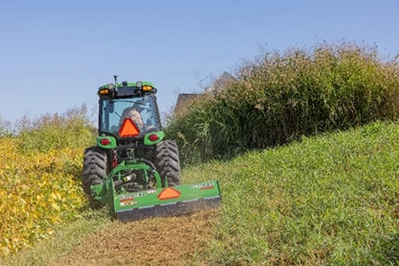 Rear view of a green tractor operating a flail mowing attachment on a grassy hillside, cutting a path through tall vegetation and weeds under a clear blue sky