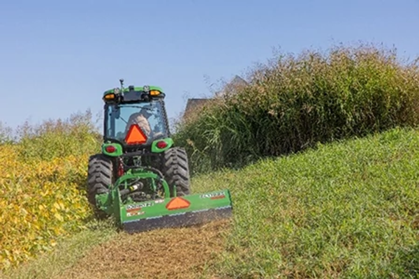Rear view of a green tractor operating a flail mowing attachment on a grassy hillside, cutting a path through tall vegetation and weeds under a clear blue sky