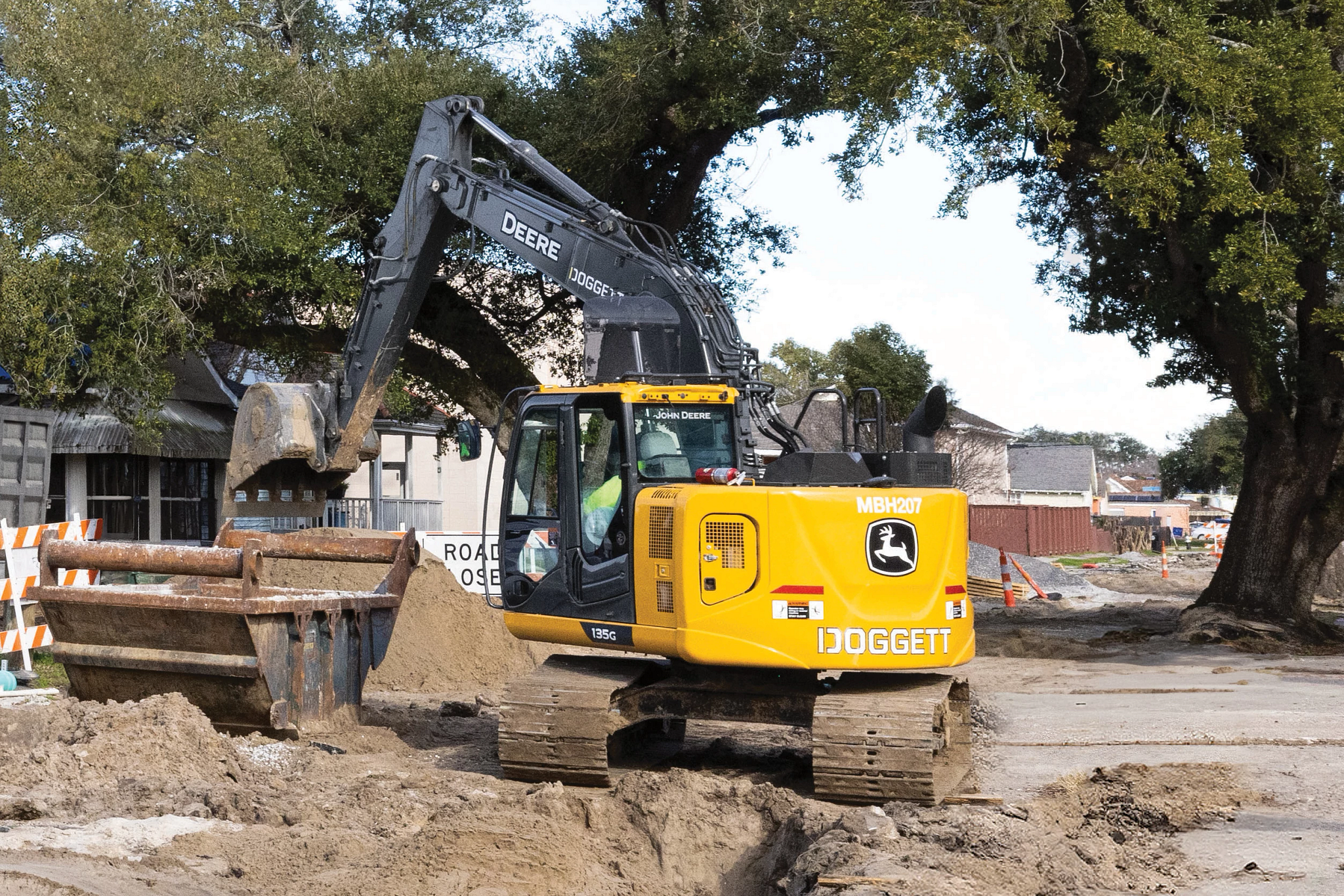 Yellow John Deere excavator at a roadside construction site, with its arm extended over a bucket near a trench and dirt pile, surrounded by barriers, trees, and nearby buildings