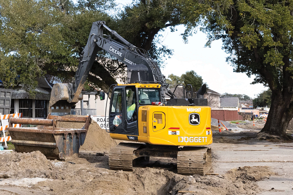 Yellow John Deere excavator at a roadside construction site, with its arm extended over a bucket near a trench and dirt pile, surrounded by barriers, trees, and nearby buildings