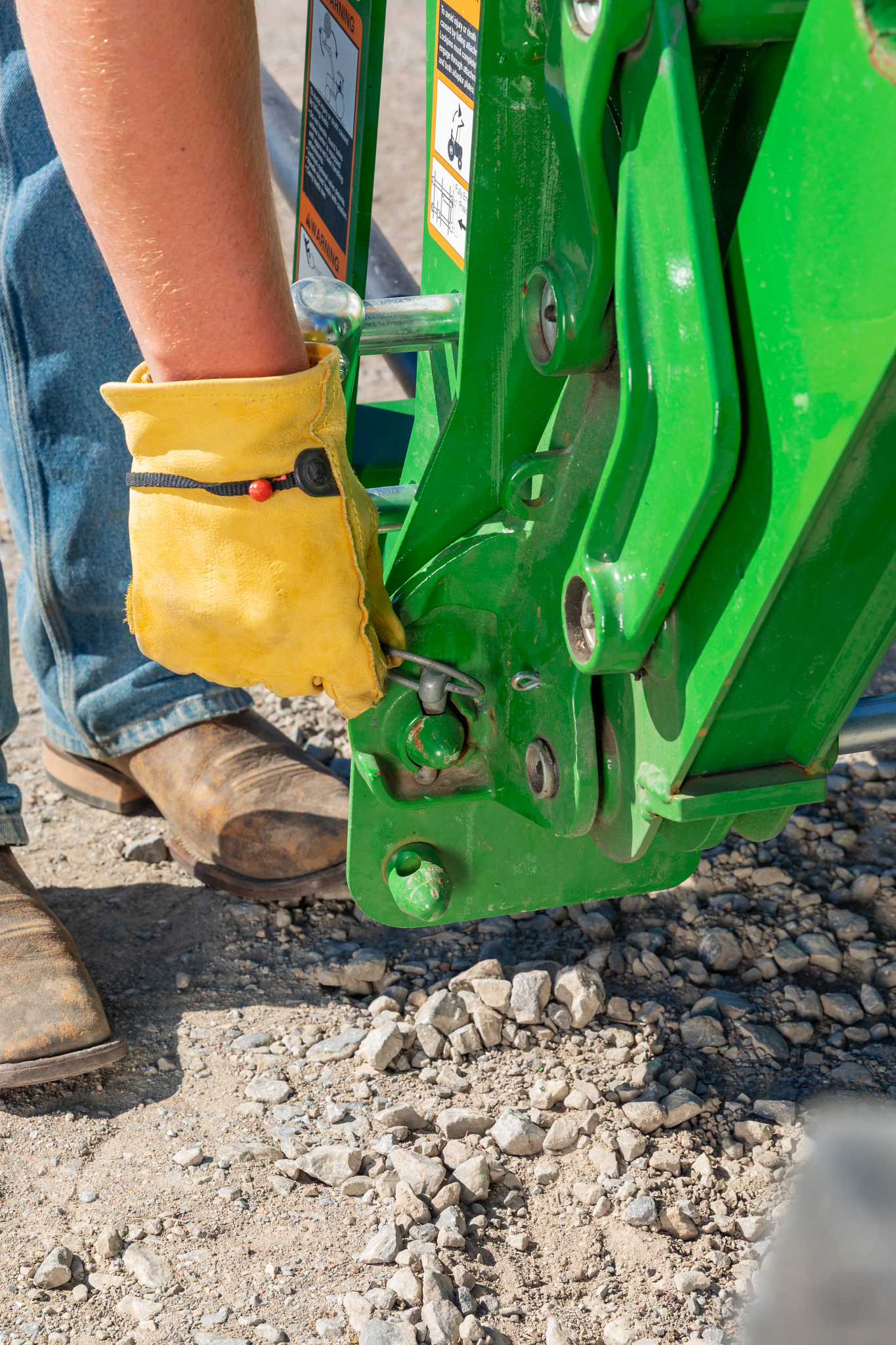 Close-up of hand securing a locking pin on a green tractor attachment, with metal linkages, safety decals, and gravel ground visible below.