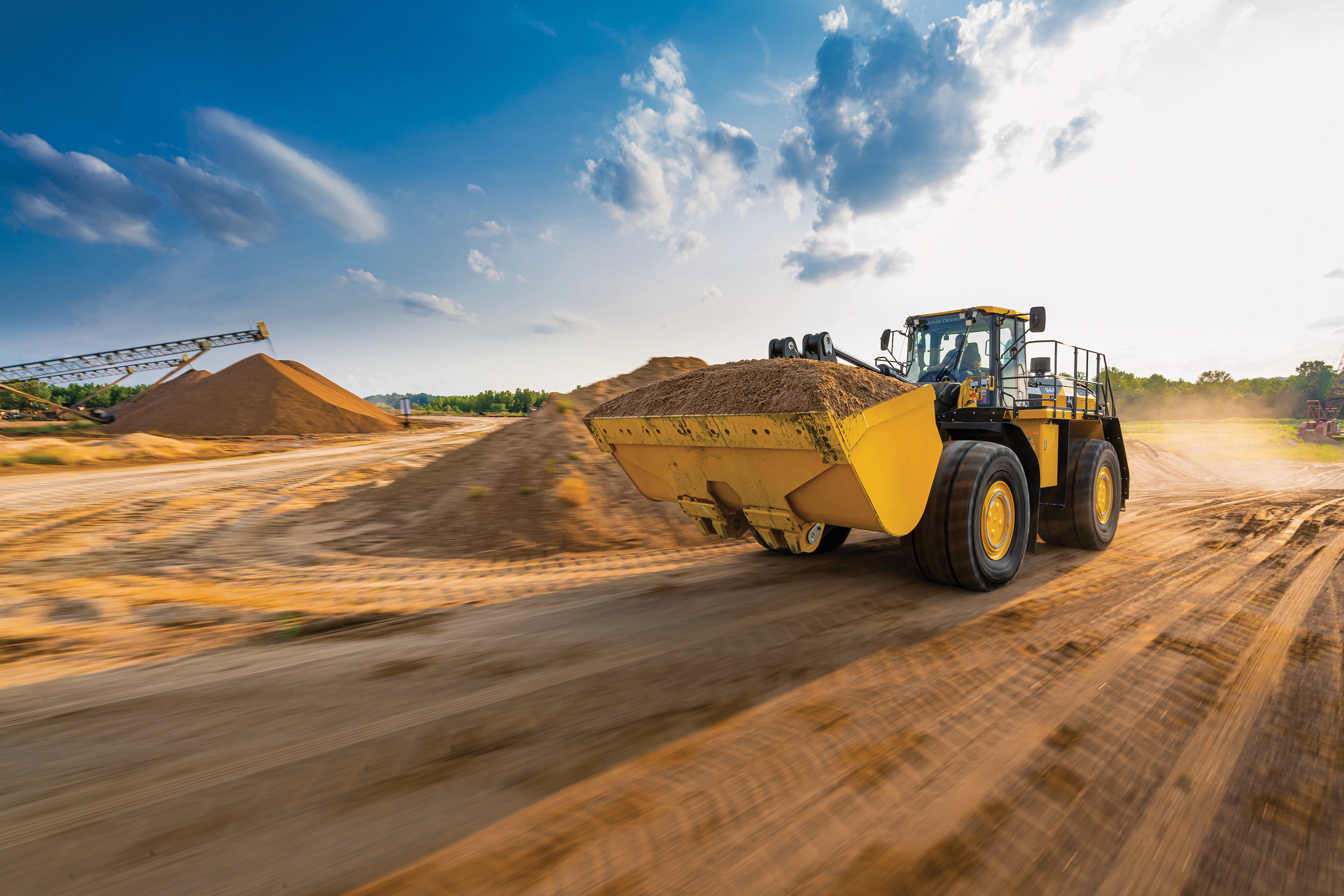 A large yellow wheel loader drives quickly along a dirt road at a quarry, carrying a full bucket of sand