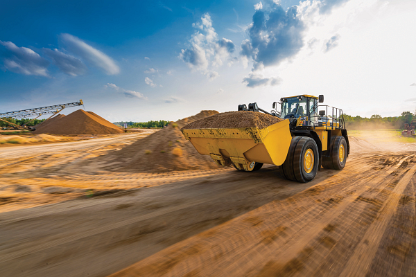 A large yellow wheel loader drives quickly along a dirt road at a quarry, carrying a full bucket of sand
