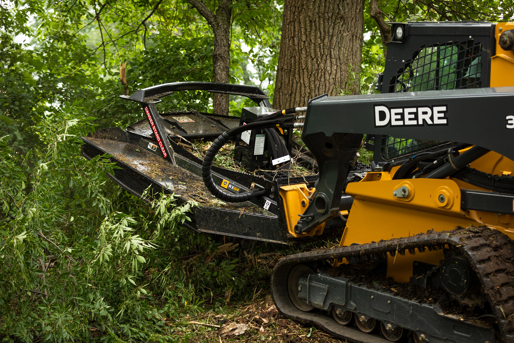 Tracked machine using a front-mounted brush cutter to clear branches and vegetation in a wooded area, with cut limbs and leaves visible around the attachment.