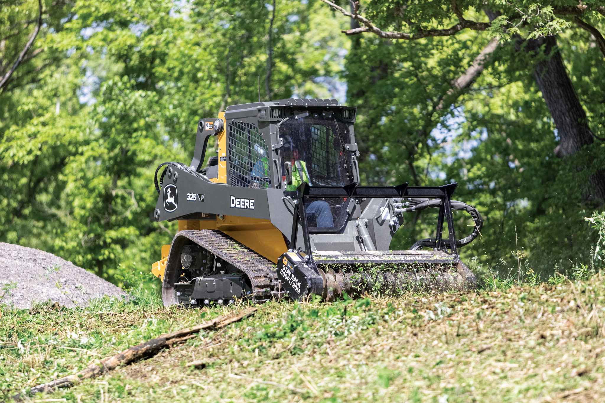 Tracked John Deere compact track loader with a front-mounted mulching attachment clearing vegetation on a grassy slope, surrounded by dense green trees in daylight.