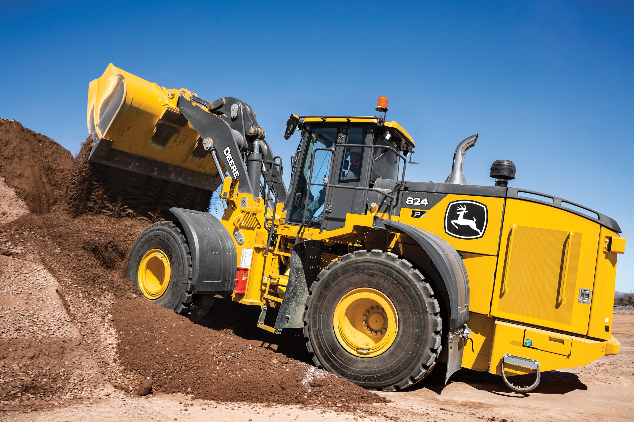 A large yellow front-end loader operates on a dirt slope, using its raised bucket to move and spread reddish soil