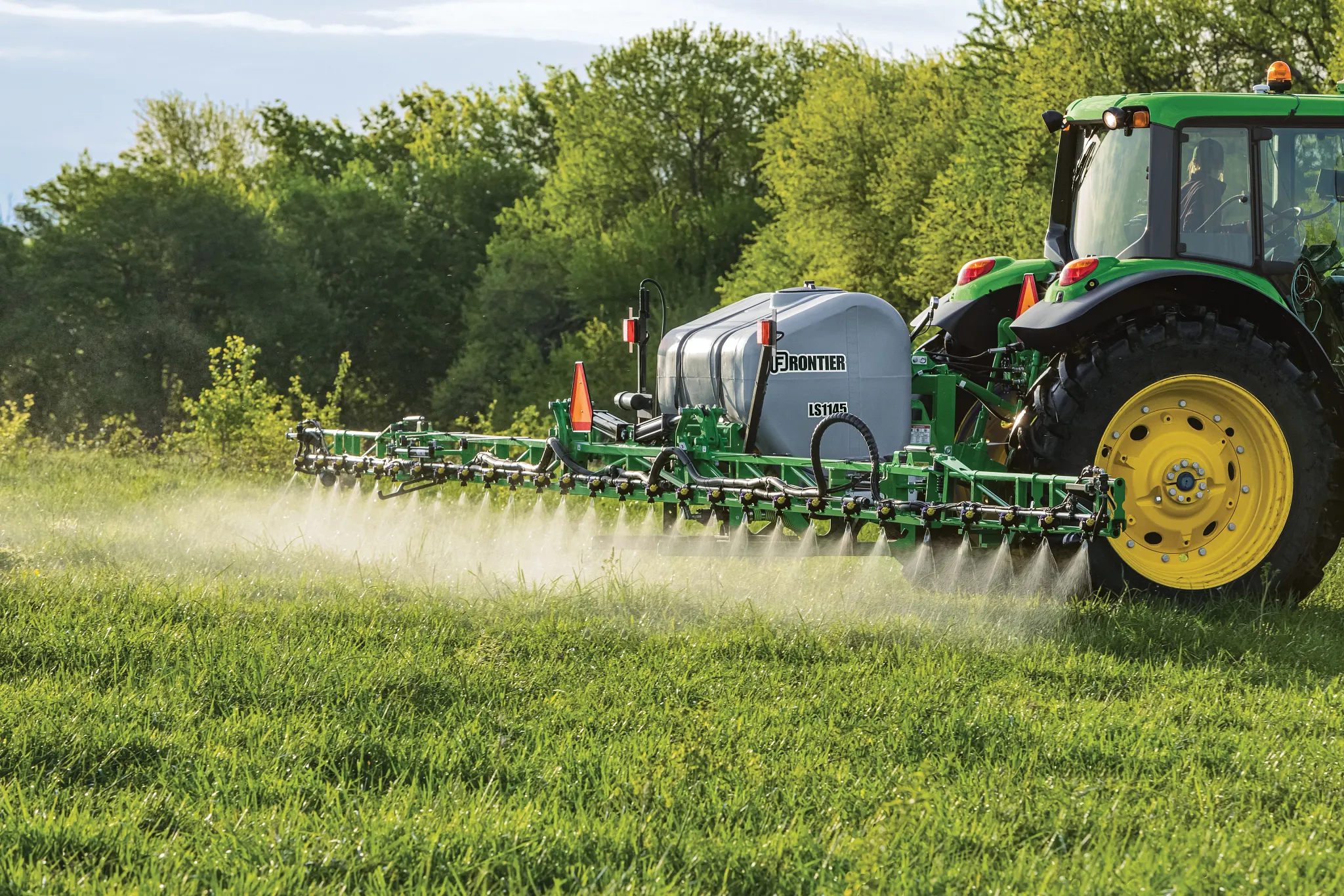 A tractor pulls a wide boom sprayer, misting crops across a grassy field, with trees in the background under daylight conditions